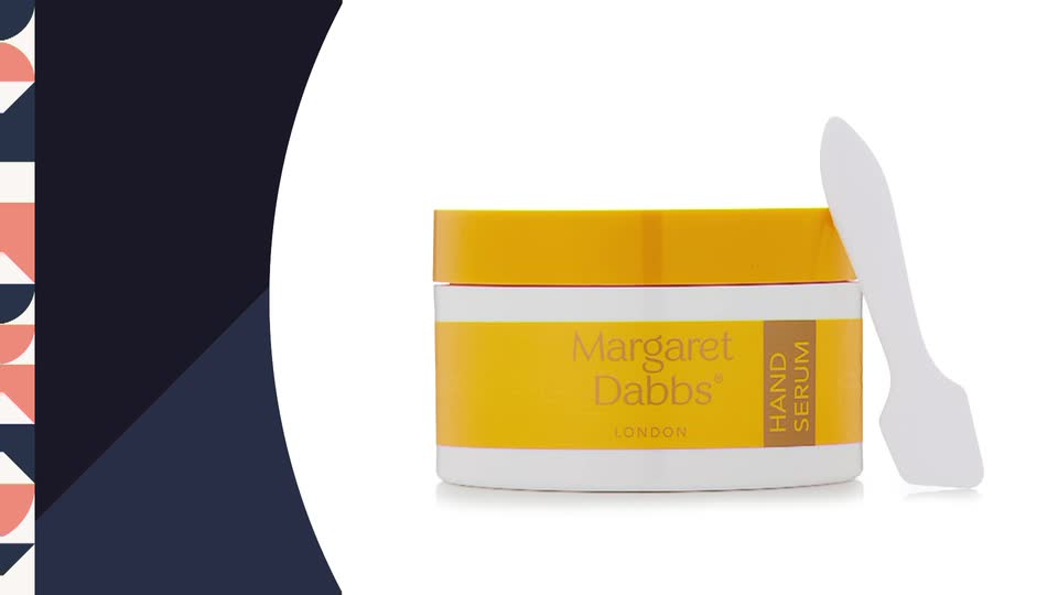 A jar of Margaret Dabbs hand serum sits on a white surface, its yellow lid and label contrasting with the white container. Beside it, a small, white spatula leans, ready for use.
