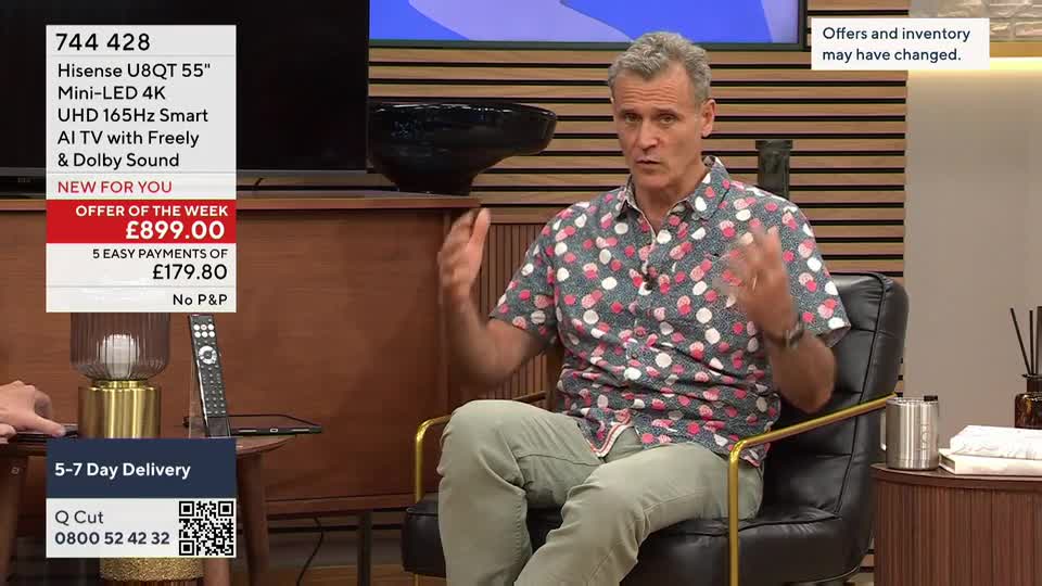 A man in a patterned shirt gestures with his hands, seated on a black chair, as a deal for a television is advertised on QVC. The screen displays the price and payment options for the "Offer of the Week."
