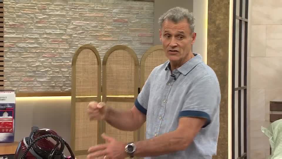 The man in the blue shirt gestures emphatically, his hands moving to emphasize a point. A red and black iron sits on the table in front of him, next to a product box.
