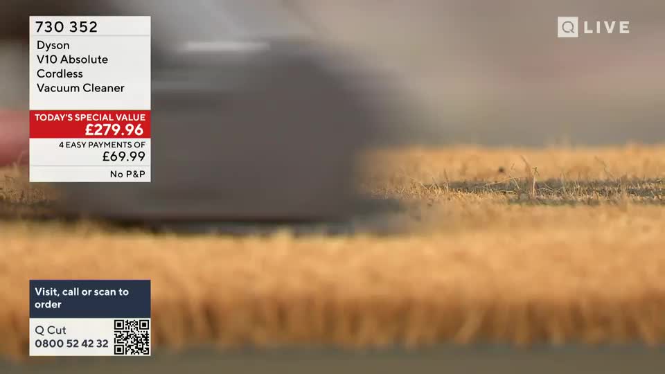 A vacuum cleaner, likely a Dyson, is being demonstrated on a rug during a QVC UK broadcast. The machine is moving across the textured surface, and the price is displayed on screen.
