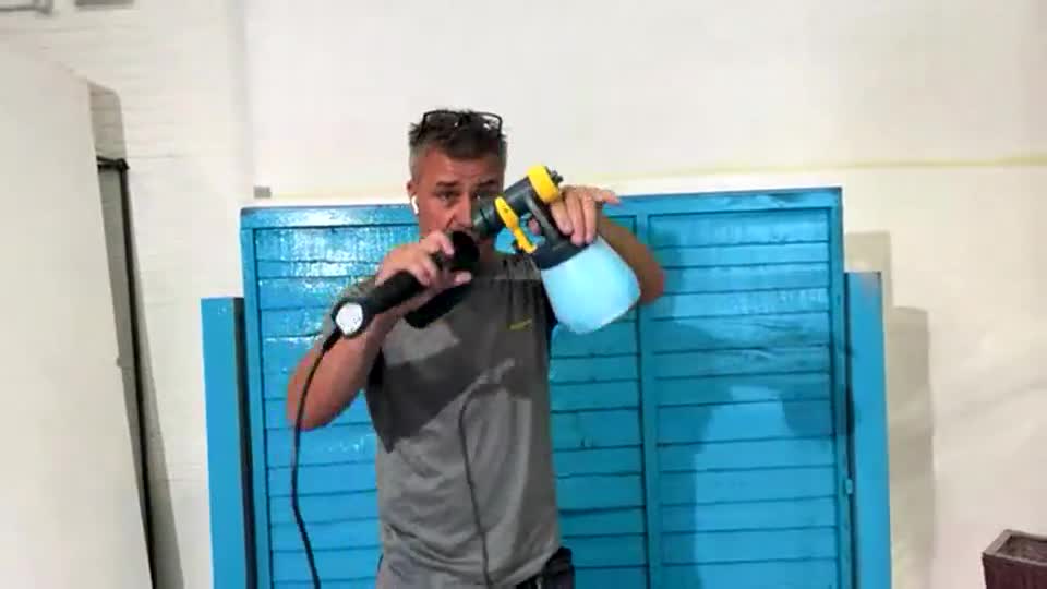 The man in the grey shirt is holding a microphone and a spray gun, demonstrating its use in front of a blue wooden backdrop. He's talking into the mic, likely selling the product on a QVC-style show.
