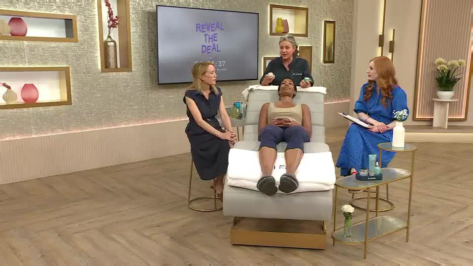 On the QVC set in the United Kingdom, a woman lies on a reclined chair while another applies something to her face. Two other women watch, one holding a clipboard and the other seated, looking on.
