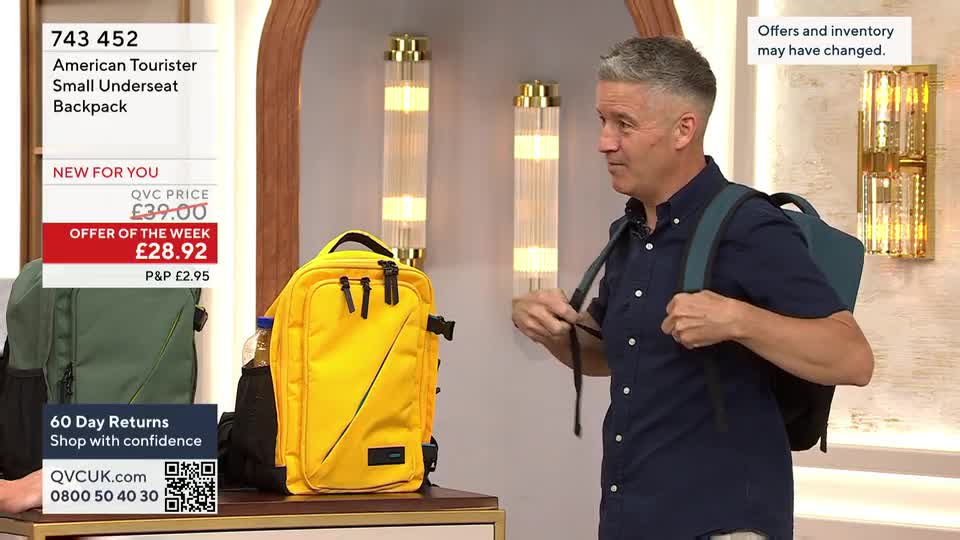 A man on QVC is demonstrating how to wear a teal backpack, adjusting the straps. The yellow and green versions of the same American Tourister bag are on display nearby.
