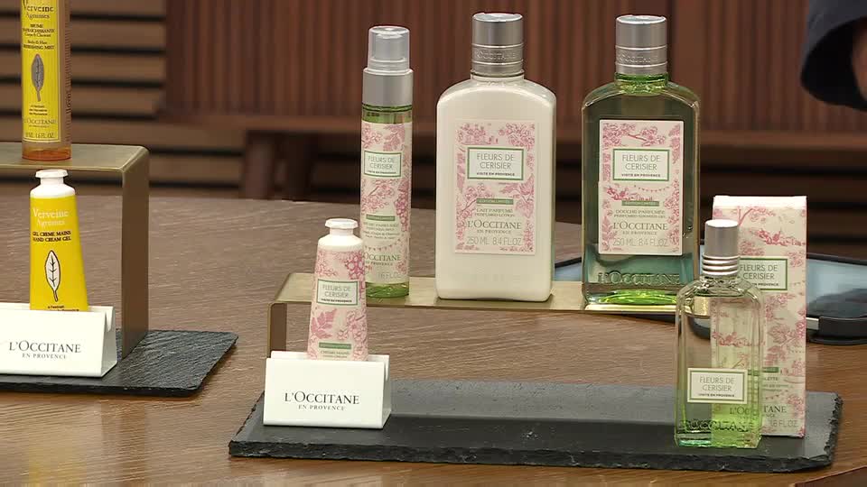 On a table, various L'Occitane products are arranged, ready for a QVC presentation. The bottles and tubes, decorated with floral designs, sit on black slate and gold-toned stands.
