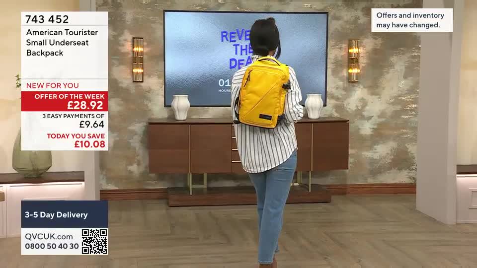 A woman in jeans and a striped shirt stands facing away, modeling a bright yellow backpack. On the screen behind her, the QVC offer of the week is displayed for an American Tourister bag.
