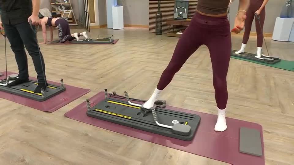 People are working out on exercise machines, each standing on a platform and holding onto a resistance cable. One woman is demonstrating a side leg lift, while others are in different positions, all using the equipment.
