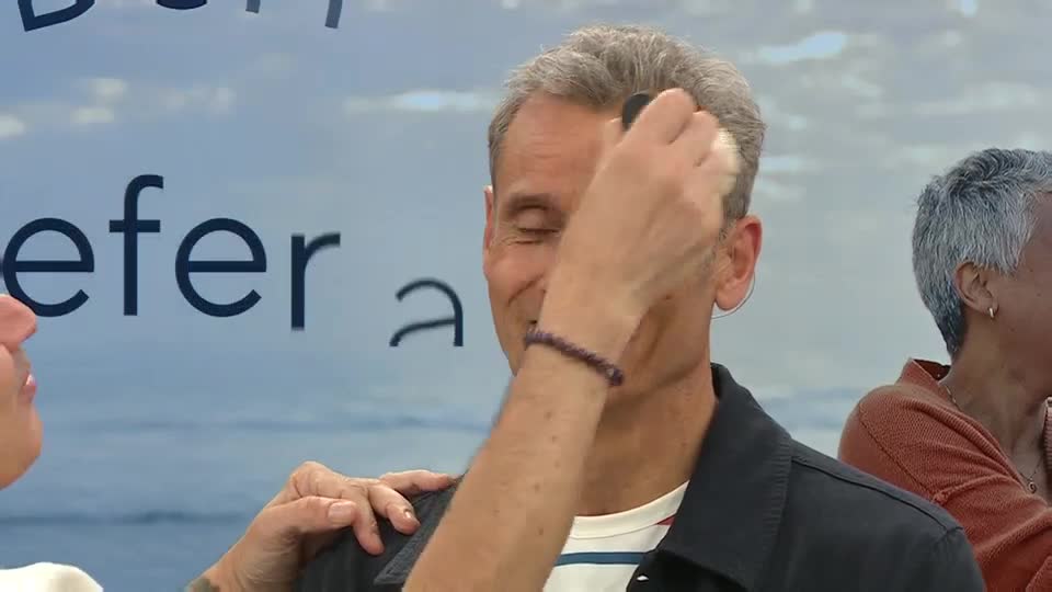 A woman is applying something to a man's forehead with a small, dark applicator. The man's eyes are closed, and he seems relaxed as he's being made up on a QVC set in the UK.
