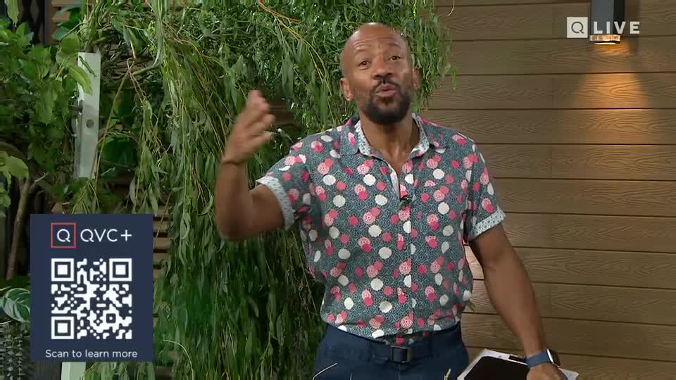 The man in the patterned shirt gestures with his hands, speaking into the camera in a UK QVC studio. Behind him, a backdrop of greenery and wood panels creates a natural setting.
