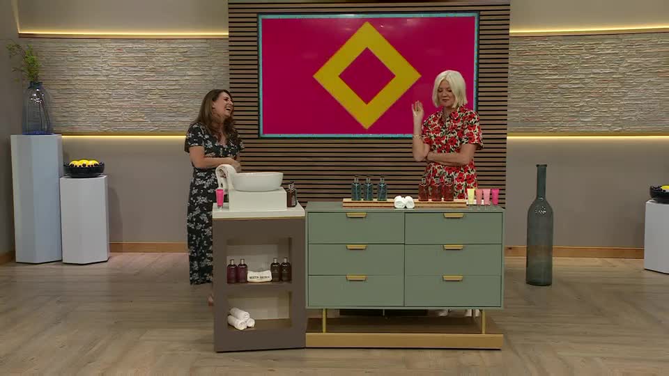 On a QVC set in the UK, one woman in a floral dress gestures with her hand while the other, also in a floral dress, laughs. A display of beauty products sits on a green cabinet.
