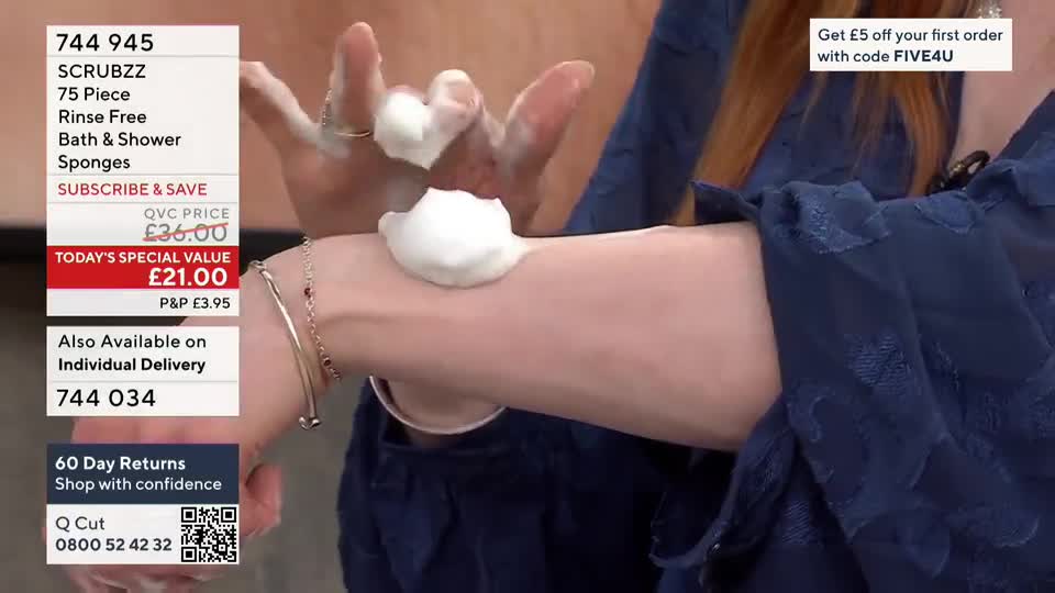 A presenter on QVC is demonstrating a bath product, applying a dollop of foamy white substance to her forearm.  The camera focuses on the product as it's gently spread across her skin.

