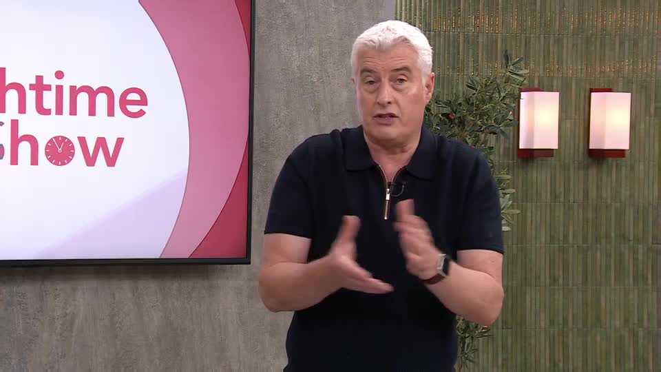 He's gesturing with his hands, explaining something on QVC's "Nighttime Show" in the UK. The host's dark polo shirt contrasts with his silver hair and the show's pink and white backdrop.
