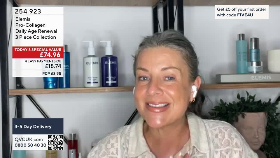 A woman with graying hair smiles directly at the camera, wearing a headset and a light-colored top. Behind her, products from Elemis are displayed on shelves, and a graphic on the screen advertises a special offer from QVC.
