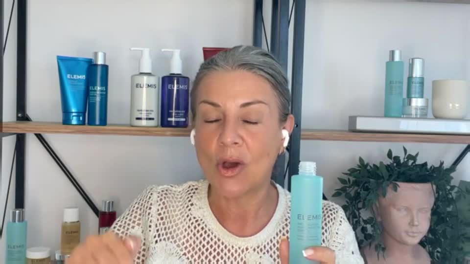 A woman, wearing earbuds, holds a turquoise bottle, gesturing with her other hand as she speaks. Behind her, on shelves, are more bottles and jars of skincare products, likely for a QVC segment in the UK.
