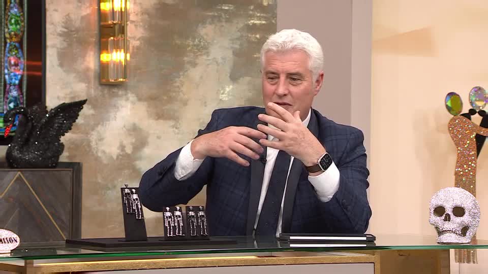 A man in a suit gestures with his hands, likely explaining something to the audience. On a display table, a few pairs of earrings are arranged in front of him.
