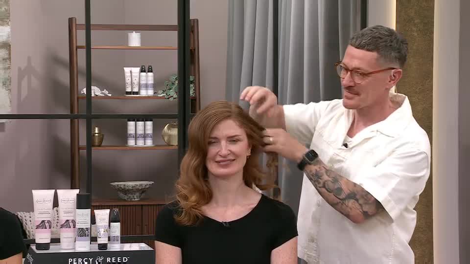 A man in glasses and a white shirt is working on a woman's wavy red hair. She's smiling, and a display of hair products sits in front of them, likely for a QVC segment.
