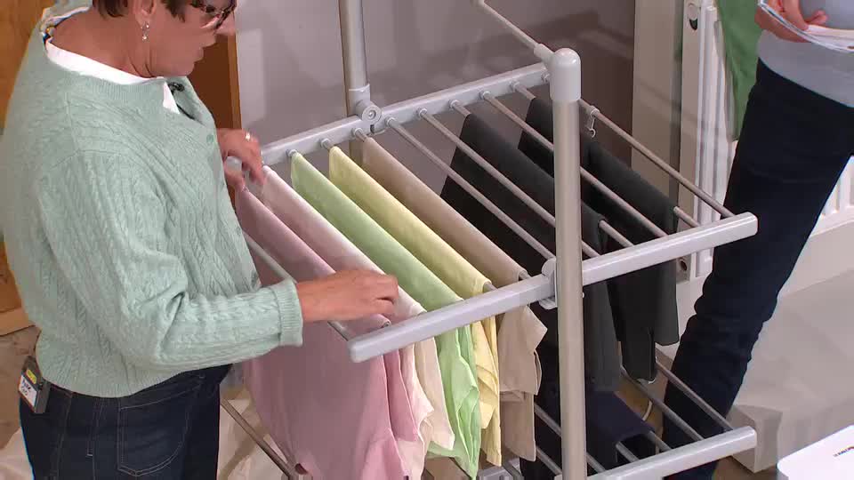 On a QVC set in the UK, a woman in a light green cardigan arranges colorful trousers on a drying rack. Another person, out of focus, holds a piece of paper, likely a script or product information.
