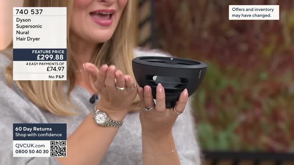 A woman on QVC in the United Kingdom holds a black hair dryer attachment, demonstrating its shape and features. She gestures with her hands, likely explaining the product's benefits to viewers.
