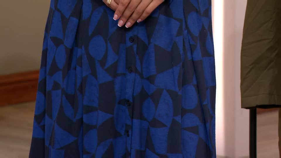 A woman's hands, adorned with rings, rest above a buttoned dress patterned with blue and black geometric shapes. The dress is on display, likely for a QVC segment from the United Kingdom.
