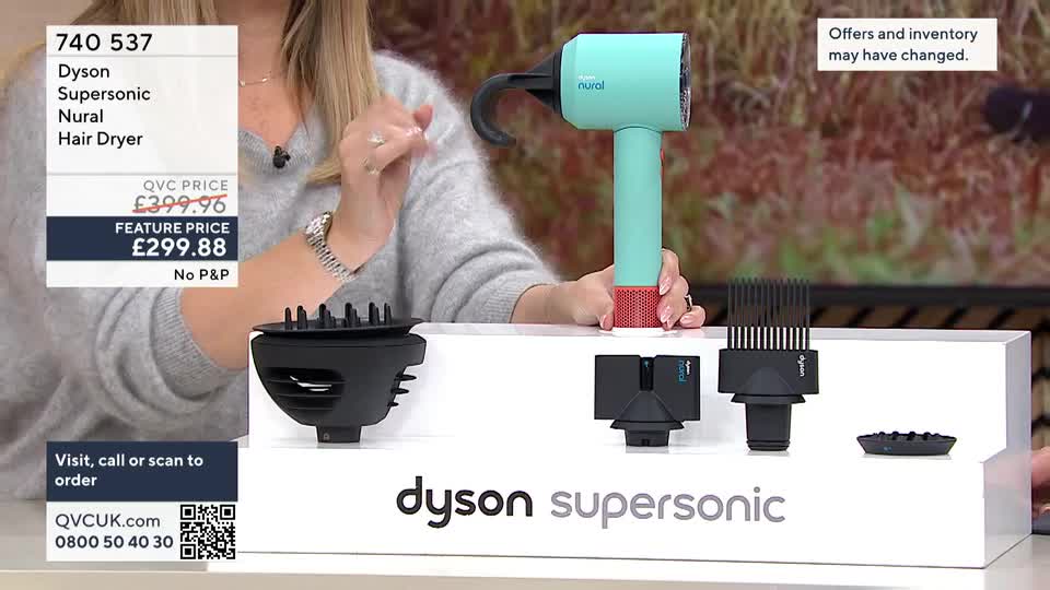 On a QVC set in the UK, a woman holds a teal and red Dyson Supersonic hair dryer, displaying its various attachments. The price is clearly visible, and the camera focuses on the product's features.
