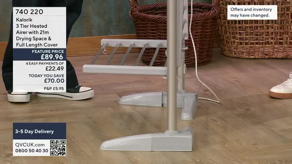 A heated airer, the focus of a QVC UK presentation, stands on a wooden floor. The product's price and details are displayed on a graphic next to a person's legs and feet.
