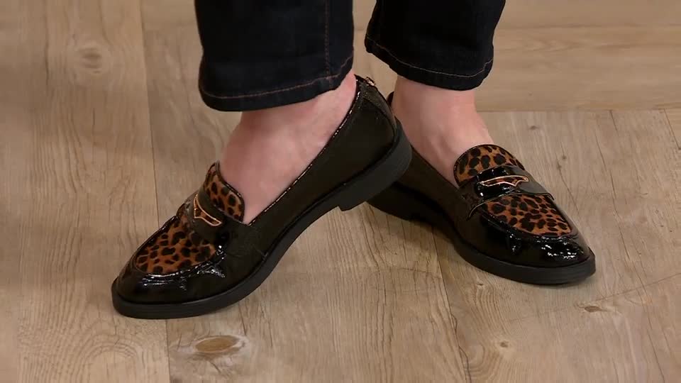 Black patent leather loafers with leopard print accents are showcased on a wooden floor. The shoes are stationary, with the model's feet in a relaxed position.
