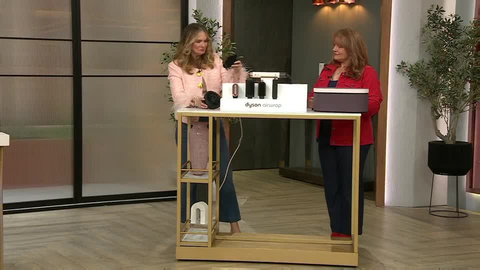 On a QVC set in the UK, a woman in a pink jacket holds up a black hair styling tool, demonstrating it to the camera. Her colleague, wearing a red jacket, stands nearby, looking on at the product display.
