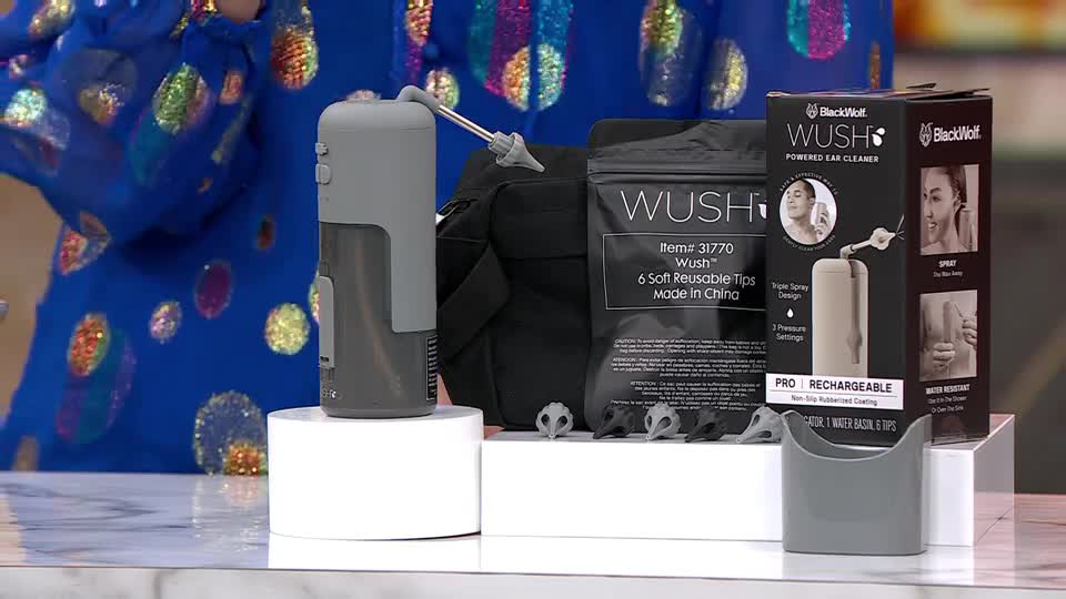 A grey ear cleaning device and accessories are arranged on a white platform, with a presenter in a sequined blue top standing nearby. The QVC studio lighting highlights the product packaging, including a box labeled "Wush."
