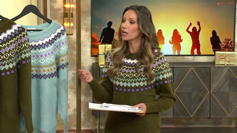 The woman in a patterned sweater gestures towards a light blue knit hanging on a rack. Behind her, a screen shows silhouettes of people dancing against a warm, sunset-like backdrop.
