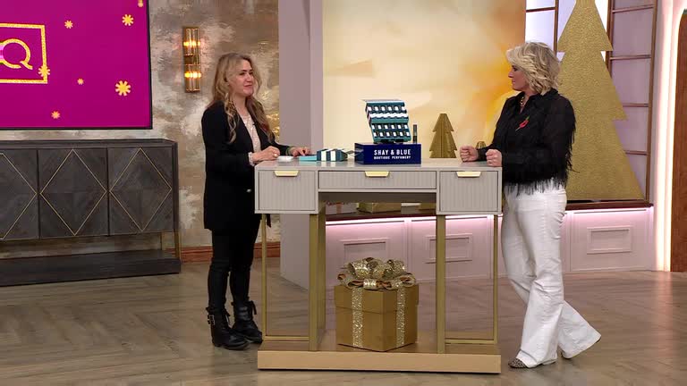 Two women stand by a desk, presumably on a QVC set in the United Kingdom, with a display of gift boxes and perfume. One gestures towards the camera as the other looks on, both framed by festive decorations.
Two women stand by a desk, presumably on a QVC set in the United Kingdom, with a display of gift boxes and perfume. One gestures towards the camera as the other looks on, both framed by festive decorations.