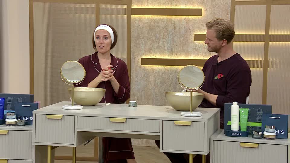 A woman in a headband and pajamas gestures with her hands, speaking to the camera. Beside her, a man in a burgundy sweater looks on, both seated at a vanity table with skincare products displayed.
