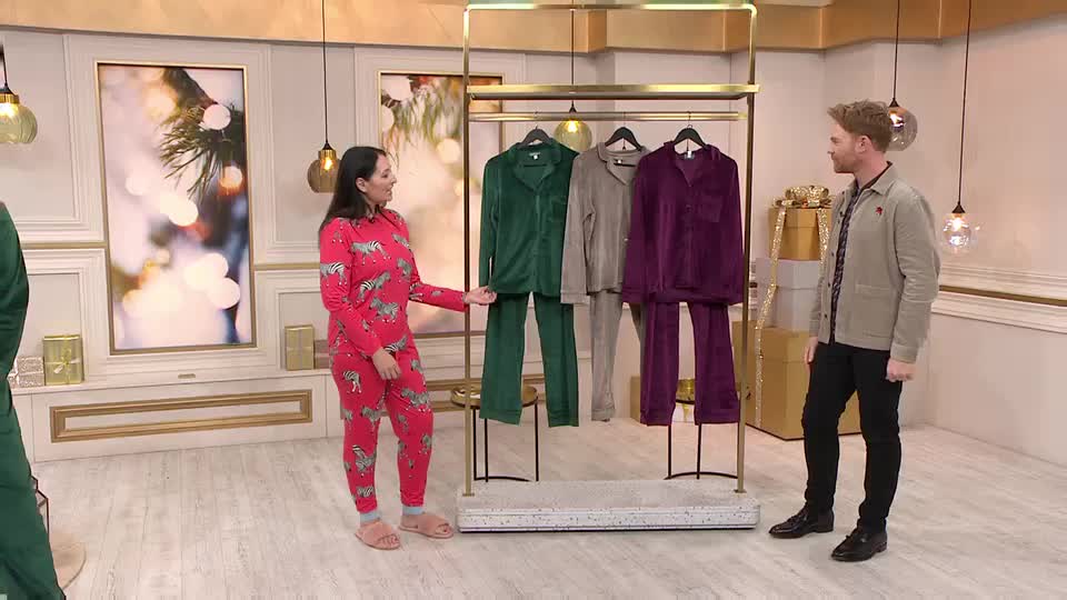 On a QVC set in the UK, a woman in a red zebra-print pajama set gestures toward a rack of velvet sleepwear. A man in a jacket stands nearby, looking on as she presents the garments.

