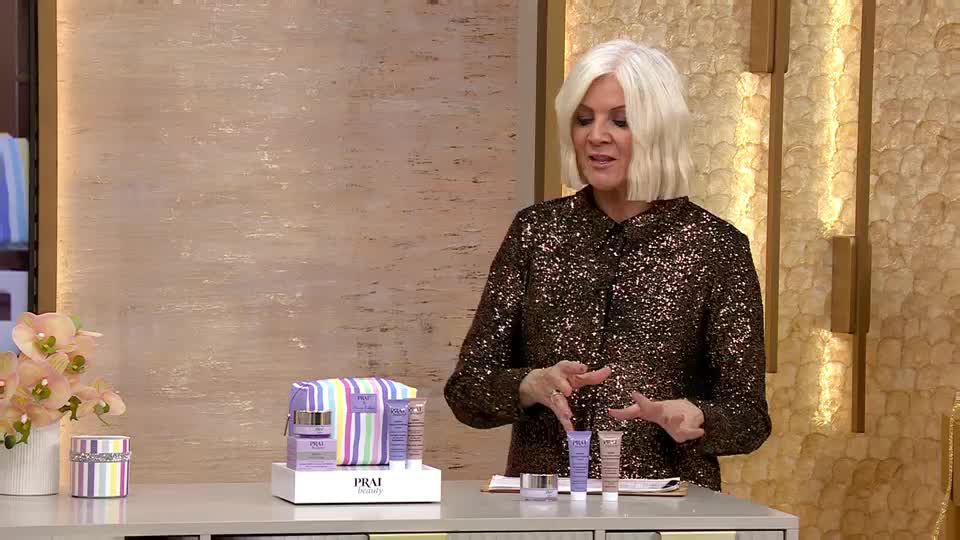 A woman in a sparkly top gestures towards beauty products, holding them up for the camera. The QVC presenter is surrounded by a display of skincare items, ready to be demonstrated.
