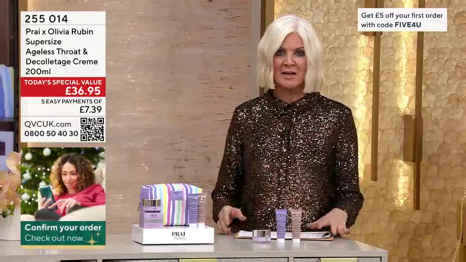A woman in a sparkly top stands behind a table, gesturing towards a selection of skincare products. On the screen, the QVC UK logo and product details are displayed, along with a promotion for a discount.

