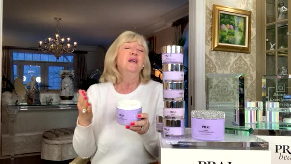 A woman in a white sweater holds a jar of lavender-colored cream, gesturing with a pink-painted nail. Behind her, a stack of similar jars sits next to a display of beauty products, likely for a QVC segment.
