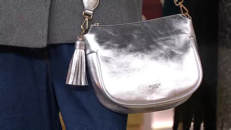 A silver Osprey London handbag is displayed, the light catching its metallic sheen. The presenter's grey top and blue jeans are a simple backdrop for the bag's detailing, including a tassel and gold chain.
