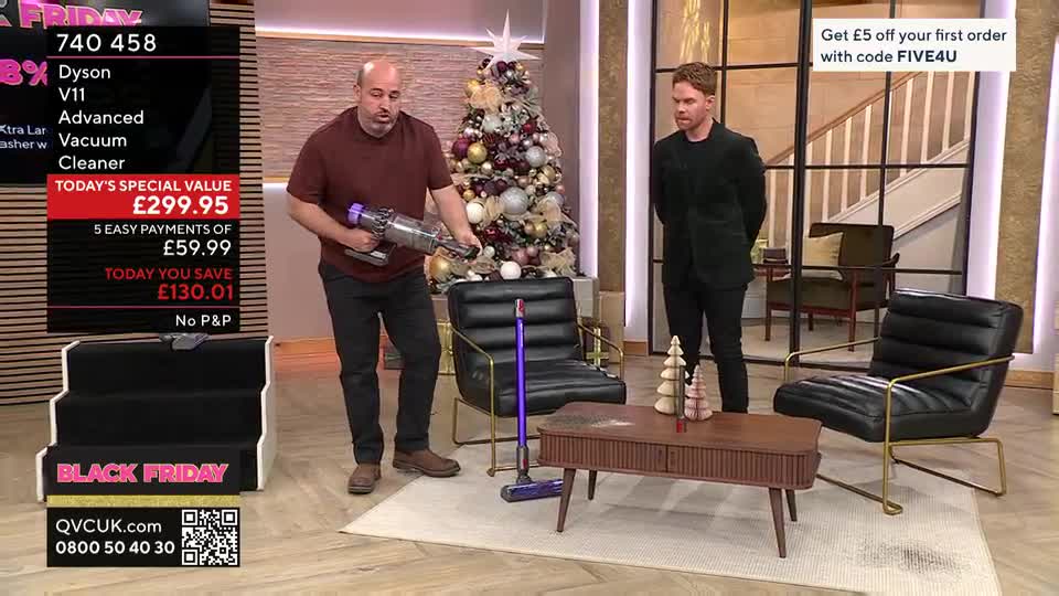 A man in a maroon shirt holds a silver and purple vacuum cleaner, demonstrating it near a chair. Across the small studio set, another man in a black suit stands, arms at his sides, as if waiting for his cue on the QVC show.

