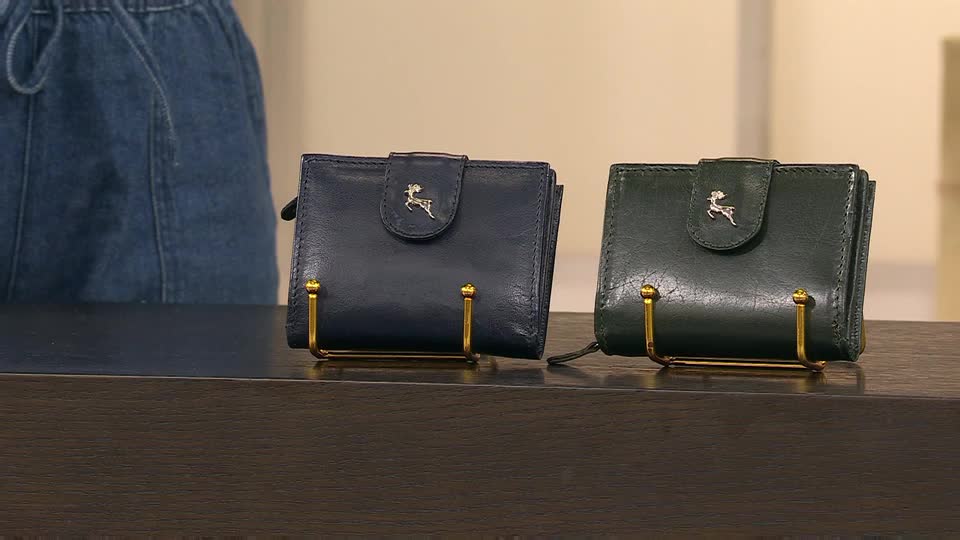 Two leather wallets, one navy and one green, sit on display, each held up by a small gold stand. A presenter in denim jeans is partially visible in the background, likely on a QVC show in the United Kingdom.
