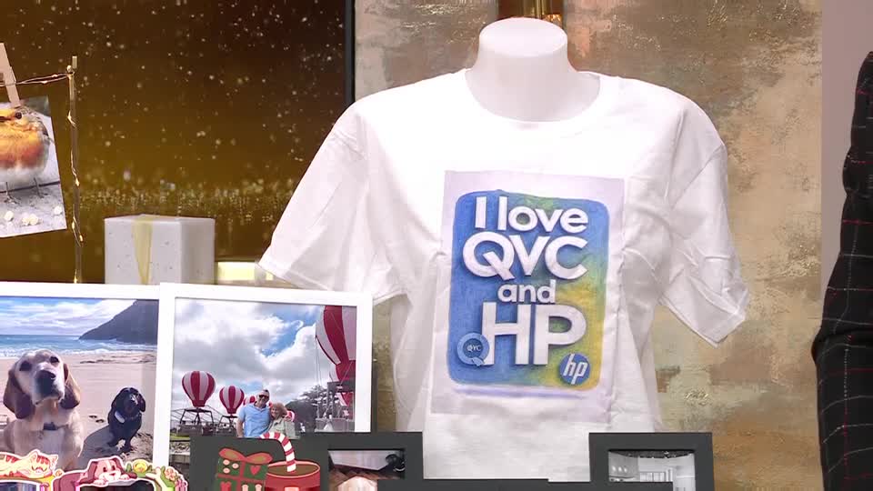 A white t-shirt on a mannequin displays a "I love QVC and HP" graphic, surrounded by framed photographs and a small gift. The background has a warm, golden glow, suggesting a studio setting.
