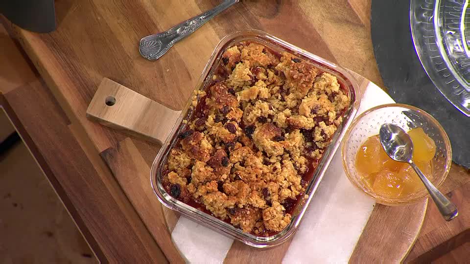 A baked crumble, golden-brown and bubbling, sits in a glass dish on a wooden board. Beside it, a small bowl holds a few glistening pieces of fruit, ready to be spooned over the dessert.
