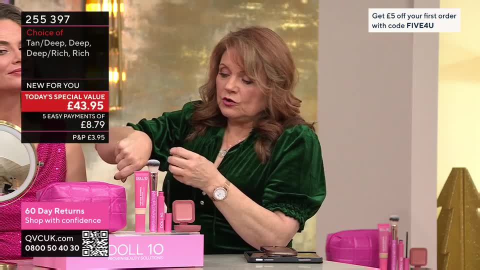 A woman in a green velvet top holds up a makeup brush, gesturing towards the camera. On a table in front of her, a pink box displays various cosmetic products, with a QVC logo visible nearby.
