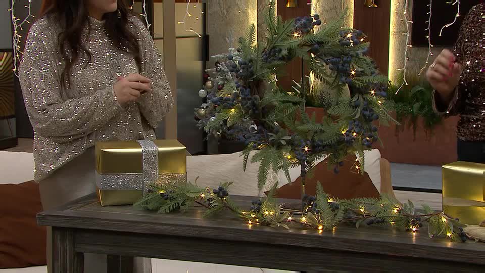 Two women are standing behind a wooden table, showcasing Christmas decorations. A wreath and garland, both adorned with berries and lights, sit atop the table alongside wrapped gifts.
