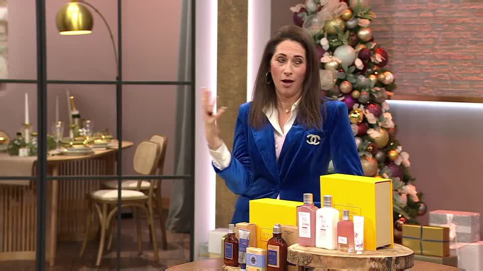 The woman in the blue velvet jacket gestures towards a table laden with gift sets, likely for a QVC segment. Behind her, a Christmas tree twinkles, and a table is set for a festive meal.
