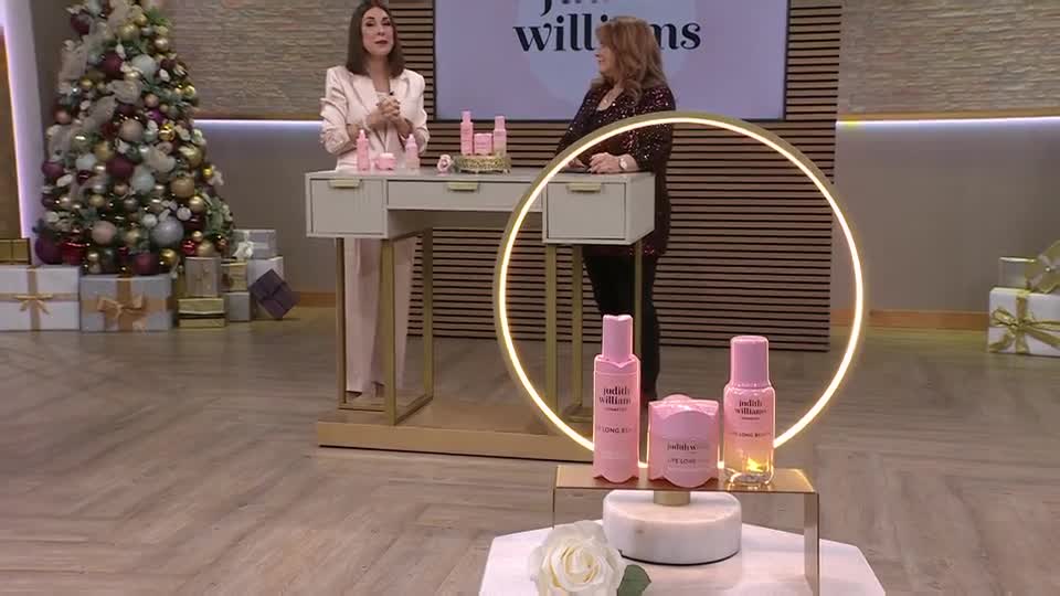 Two women stand in a studio set, one in a pink suit, the other in a sparkly top. They're presenting a line of pink skincare products, with a Christmas tree and wrapped gifts in the background.
