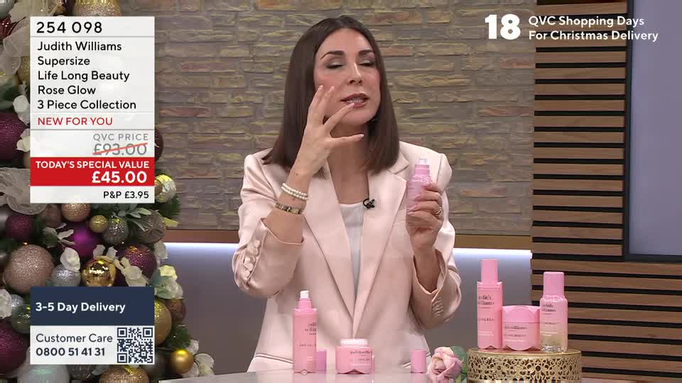 A woman in a pink blazer is demonstrating a skincare product, dabbing it on her cheek. Several matching pink bottles and jars sit on a small display table in front of her.
