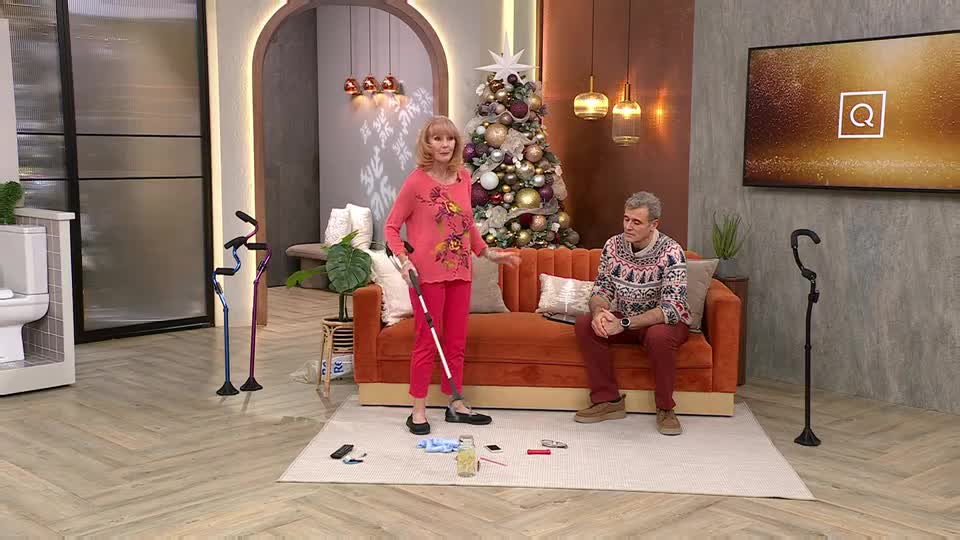 A woman in pink gestures with a white cane, while a man in a festive sweater sits on a sofa. Several walking sticks stand nearby, and a Christmas tree is in the background.
