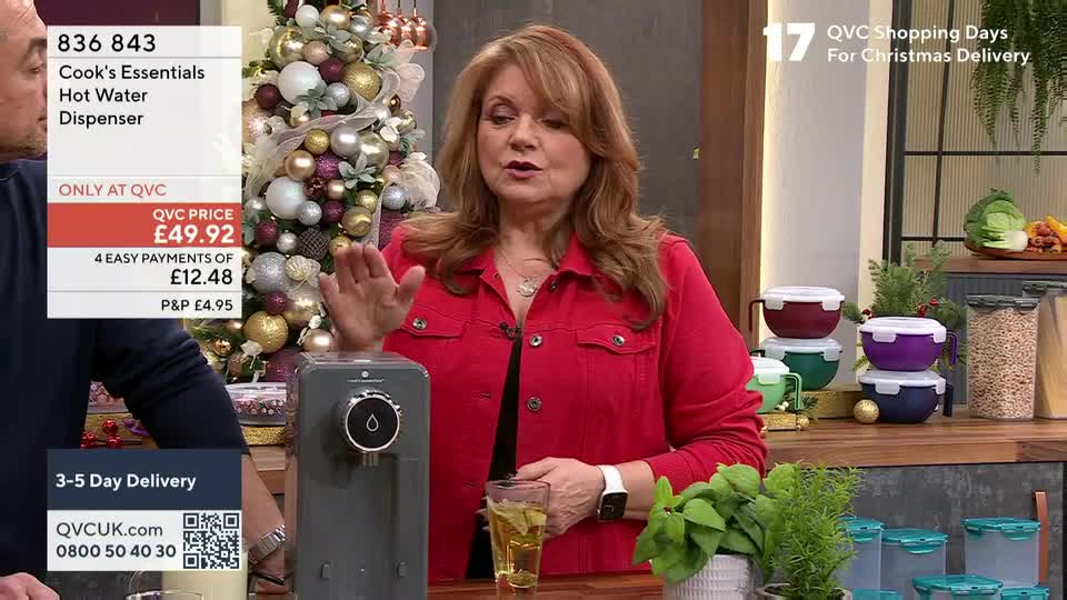 A woman in a red jacket gestures towards a small, grey hot water dispenser, holding a glass of tea. The QVC price tag for the appliance is visible next to her.
