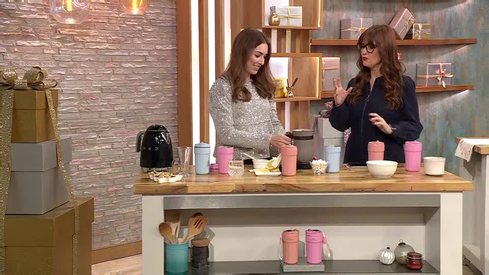 Two women stand behind a counter, one in a sparkly top stirring something into a pink travel mug. The other gestures with her hands, seemingly explaining the product on display, while a selection of mugs and snacks sit arranged on the countertop.
Two women stand behind a counter, one in a sparkly top stirring something into a pink travel mug. The other gestures with her hands, seemingly explaining the product on display, while a selection of mugs and snacks sit arranged on the countertop.