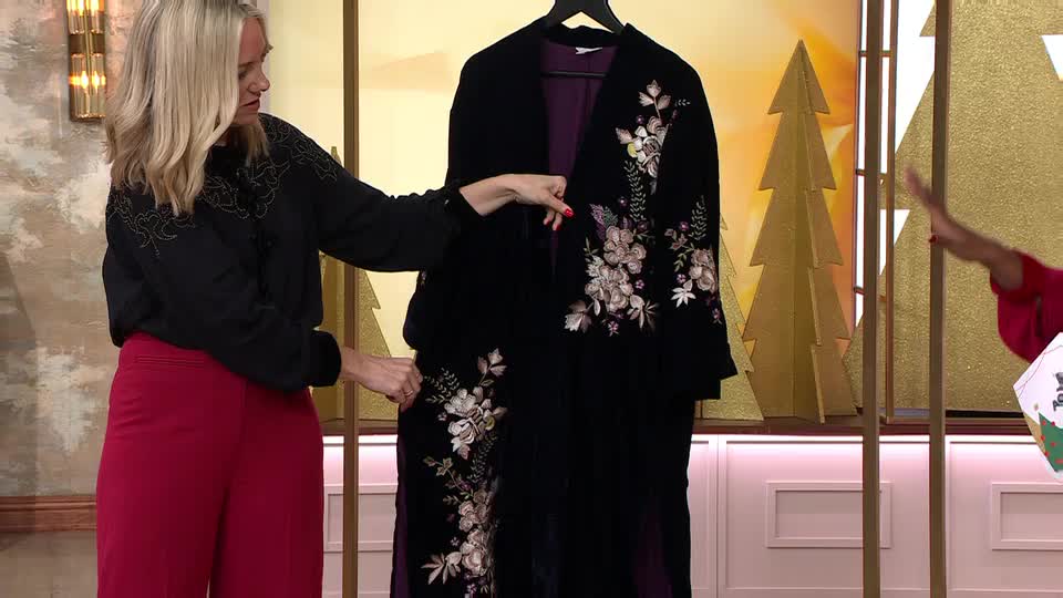 A woman in red trousers points at a long, black velvet coat with floral embroidery on QVC in the United Kingdom. The coat hangs from a rack, and another person is gesturing beside a festive backdrop.
A woman in red trousers points at a long, black velvet coat with floral embroidery on QVC in the United Kingdom. The coat hangs from a rack, and another person is gesturing beside a festive backdrop.