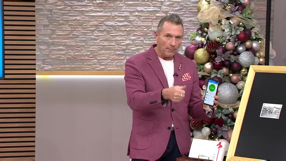 A man in a magenta blazer gestures with his right hand while holding a phone, pointing at the screen. Behind him, a Christmas tree glitters, and a small chalkboard displays a photo.
A man in a magenta blazer gestures with his right hand while holding a phone, pointing at the screen. Behind him, a Christmas tree glitters, and a small chalkboard displays a photo.