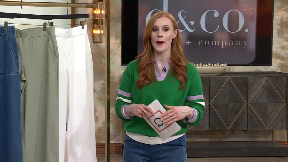 A woman in a green jumper is presenting trousers on a QVC segment. Three pairs of wide-leg trousers hang on a rail beside her.